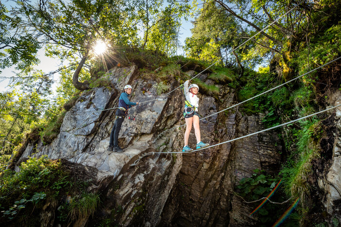 Zwei Kinder mit Helm und Klettergurt auf Seilbrücke an Felswand, sonniges Wetter, Bäume und Felsen im Hintergrund. | © Snow Space Salzburg, Christoph Huber