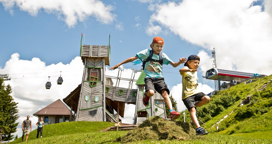 Two kids jump over a grassy mound in front of a fantasy play tower at Geisterberg adventure park in St. Johann, Salzburg | © sanktjohann.com | Mirja Geh
