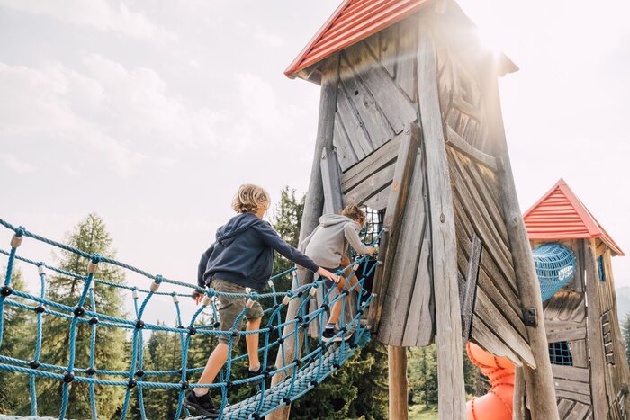 Two children climb across a blue rope bridge to a wooden play tower with red roofs in sunlight. | © JOSalzburg