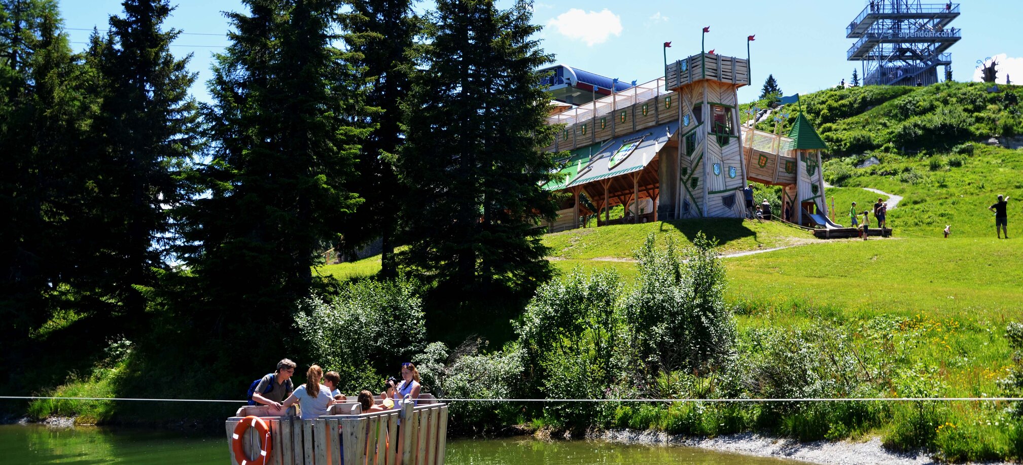 Group on wooden raft on mountain lake, behind them a castle-like play structure, trees and viewing tower visible
