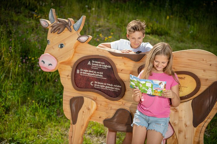 Girl reads KUHbidu booklet, boy leans on wooden cow with quiz board, green meadow and wildflowers in background | © Snow Space Salzburg, Christoph Huber