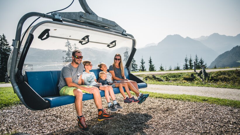 Family with three kids sitting on grounded chairlift seat, sunny weather, mountains and pine trees visible in background | © Snow Space Salzburg