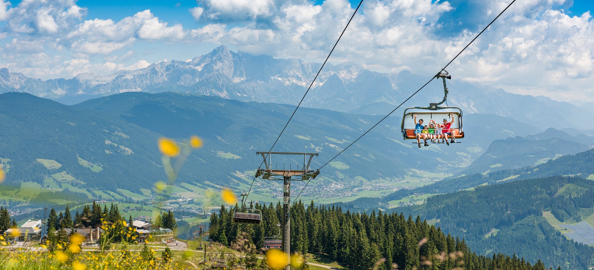 People ride chairlift over flower meadow in sunshine, mountain panorama with forests and valleys visible in background