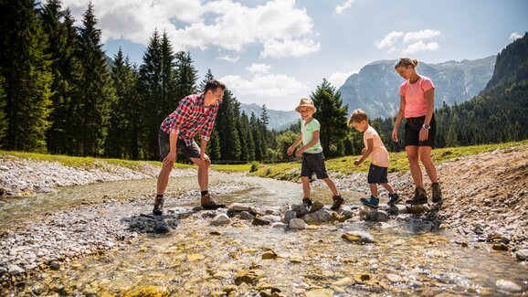 Family with two kids crossing a stream on stones, all smiling, with green forest and mountains in the sunny background