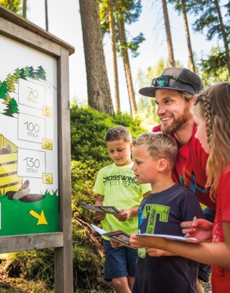 Family with three kids stands at forest quiz board with cones, all holding booklets, smiling and engaged in guessing