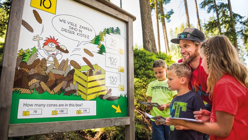Family with three kids stands at forest quiz board with cones, all holding booklets, smiling and engaged in guessing