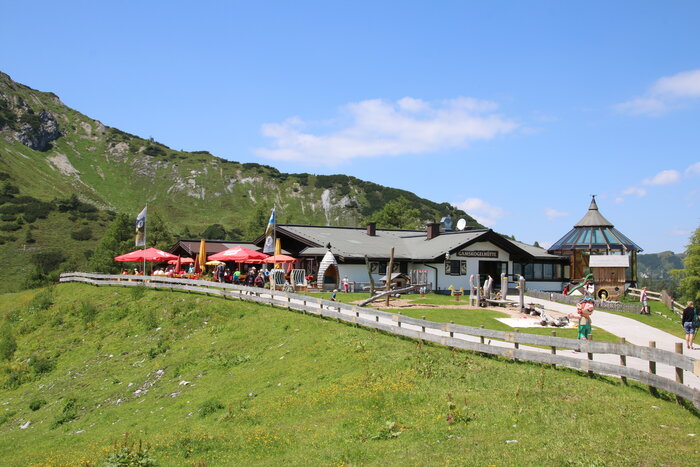 Gamskogelhütte in summer with sun umbrellas, playground and lush green mountain slopes in the background.