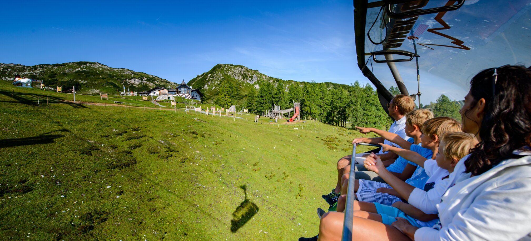 Family on summer chairlift points to mountain hut and adventure playground near Gamskogel in Zauchensee Alps.