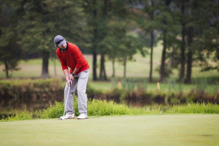 A man in a red sweater is putting on a green golf course, surrounded by trees, a pond, and calm natural surroundings.