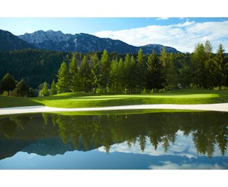 Green fairway with flag by a calm pond, surrounded by trees and backed by the Dachstein mountain range. | © GC Schladming-Dachstein