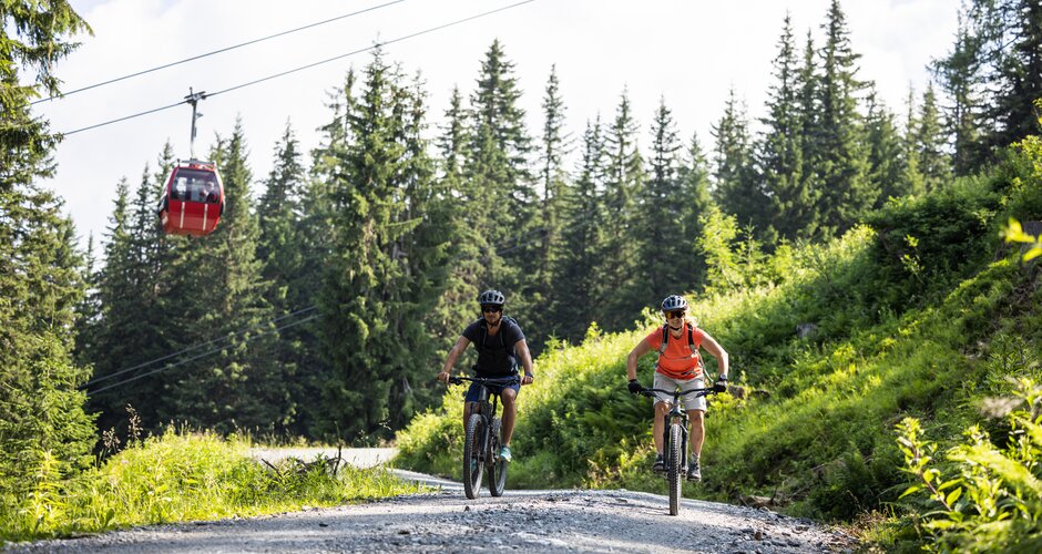 Two mountain bikers on gravel path below red gondola lift at Fulseck in lush green alpine scenery | © SalzburgerLand, Heiko Mandl