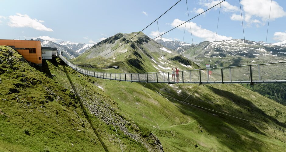 Two people stand on a suspension bridge above green alpine slopes with rocky mountains and patches of snow. | © Gasteiner Bergbahnen; Marktl Photography