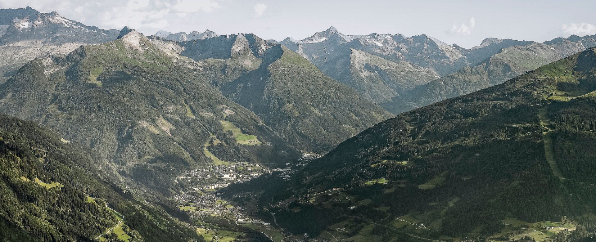 Wide view over Gastein Valley with a town below, green slopes and a long mountain range under a bright sky. | © Gasteinertal Tourismus, Marktl Photography
