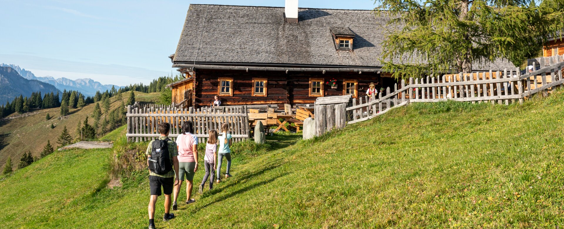 Four hikers walk uphill to a rustic alpine hut in Großarltal, surrounded by green meadows and scenic views of mountain peaks | © Lorenz Masser