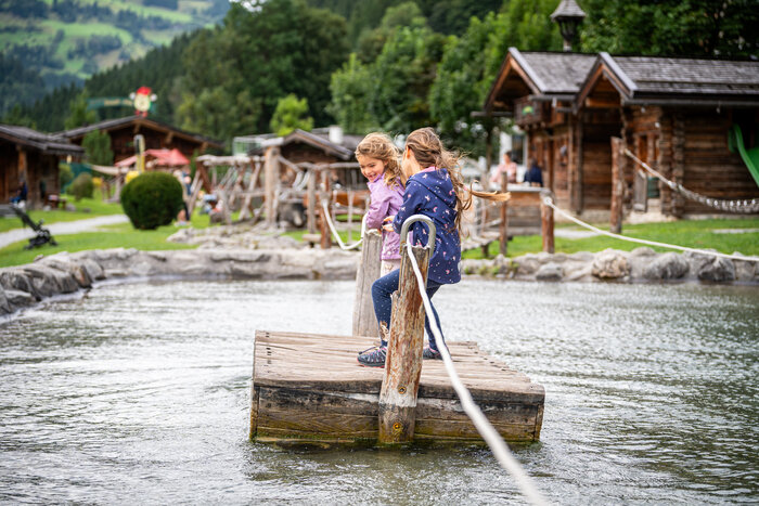 Two children play on wooden raft in water at playground in Grossarltal with huts meadow and trees | © Gipfelfieber
