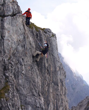 Two climbers on steep rock wall secured with rope, one ascends while the other secures; clouds and alpine peaks in background
