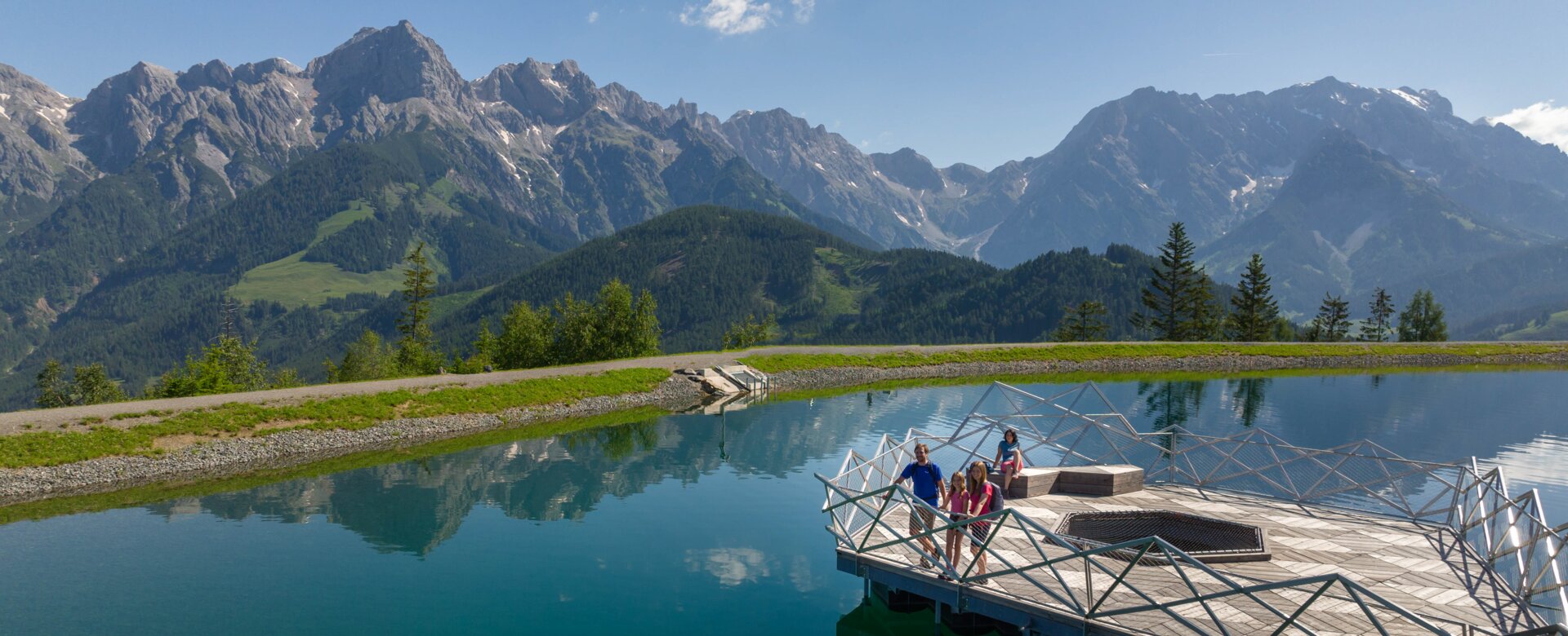 Family on wooden platform at Prinzensee in Maria Alm, clear lake water and Hochkönig mountains in background | © Roland Haschka