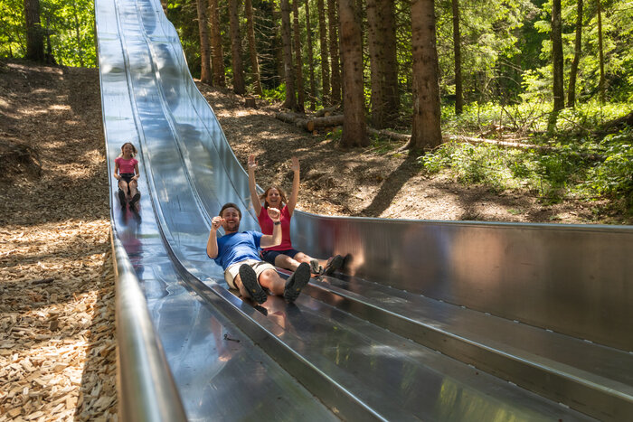 Family laughing while sliding down wide metal slide in forest slide park at Maria Alm's Natrun mountain | © Roland Haschka
