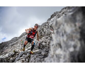 Athlete runs on rocky mountain trail in race gear with bib number, rugged terrain and another runner in the background