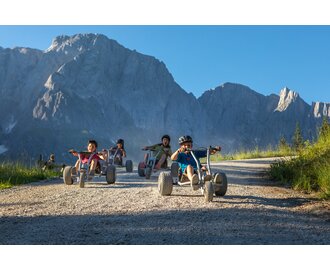Kids race downhill on mountain carts with dramatic Hochkönig peaks behind, wearing helmets, sunny day | © Roland Haschka