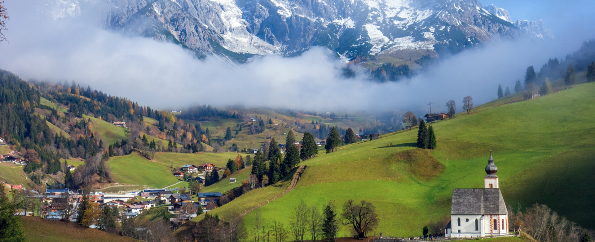 White church near Dienten on green hill, snowy Hochkönig rises above misty valley in the background