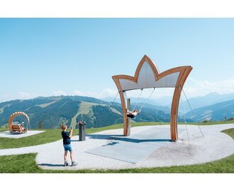 Child swings under large crown-shaped frame with Alpine view; woman takes photo; bench swing in grassy background | © Miriam Lottes