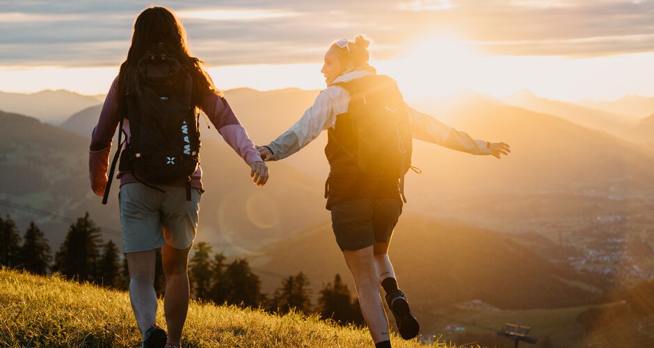 Two women with backpacks walk across a meadow in evening light overlooking mountains and valley in the Hochkönig region. | © Hochkönig Tourismus
