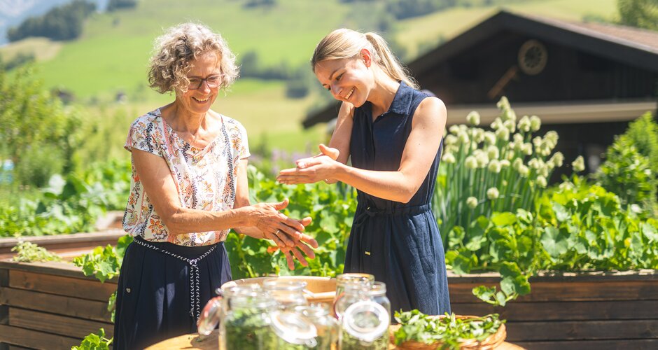 Two women examine herbs at a table with jars and a basket in front of raised beds, a house and green hills. | © Hochkönig Tourismus GmbH