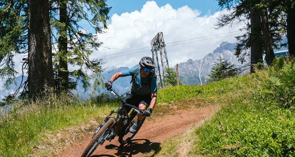 A mountain biker rides through a bend on a dirt trail between trees with mountains in the Hochkönig region. | © Hochkönig Tourismus GmbH