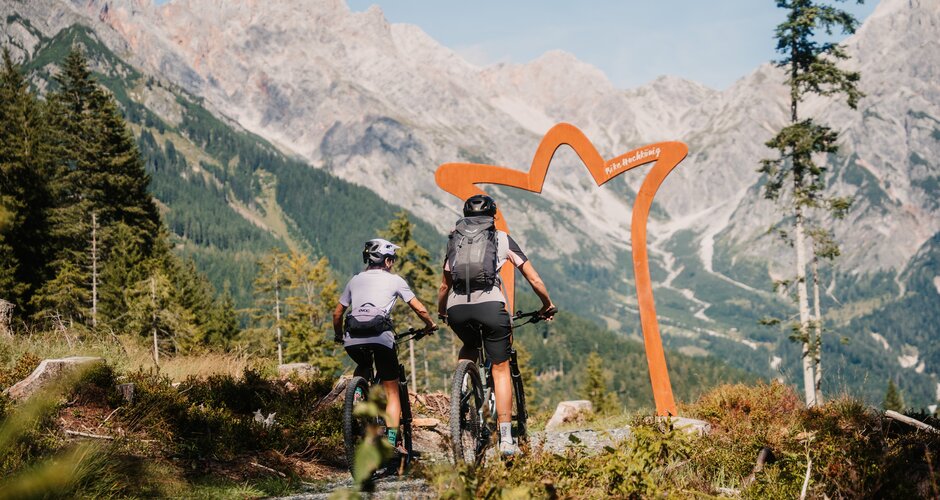 Two mountain bikers ride on a gravel trail towards an arch, with high rocky mountains in the Hochkönig region. | © Hochkönig Tourismus GmbH