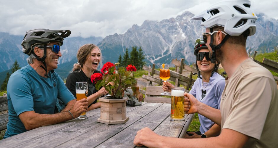 Four cyclists wearing helmets sit at a wooden table with drinks in front of mountains in the Hochkönig region. | © Hochkönig Tourismus GmbH
