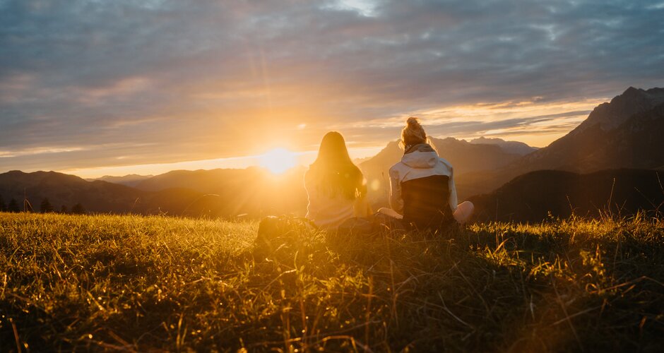 Two women sit on a grassy meadow and look towards the sunset over mountain silhouettes under a cloudy sky. | © Hochkönig Tourismus GmbH