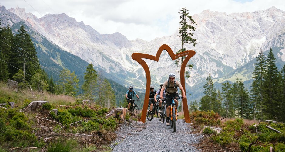 Four mountain bikers ride on a gravel path through an arch with rocky mountains and pine trees in the Hochkönig region. | © Hochkönig Tourismus GmbH