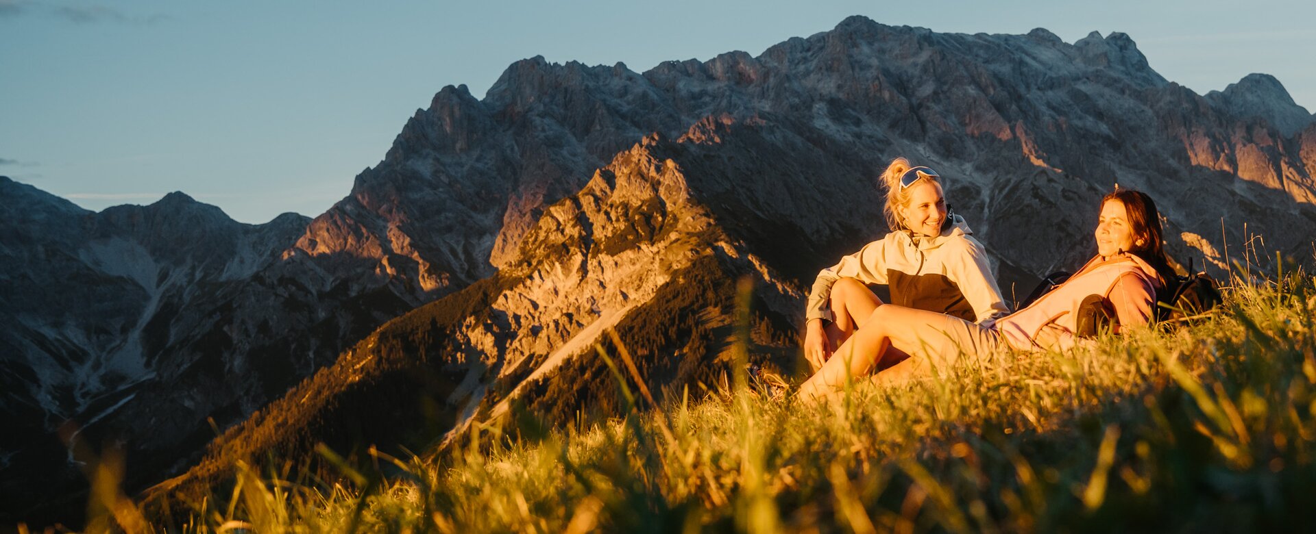 Two women sit on a grassy slope in front of a rocky mountain massif in warm evening light in the Hochkönig region. | © Hochkönig Tourismus GmbH