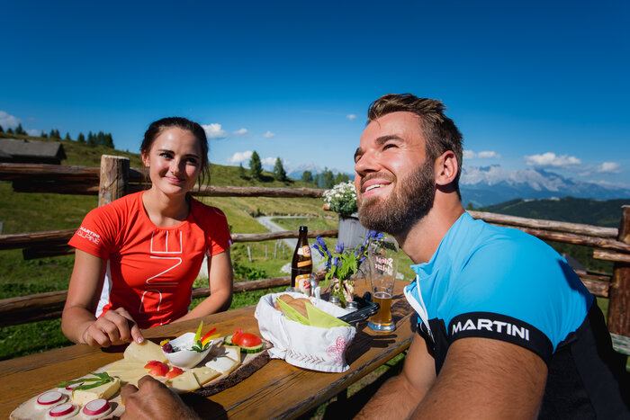 Man and woman enjoy alpine snack with cheese platter and drinks in the sun, with scenic Salzburg mountains in the background | © Salzburger Sportwelt - Christian Schartner
