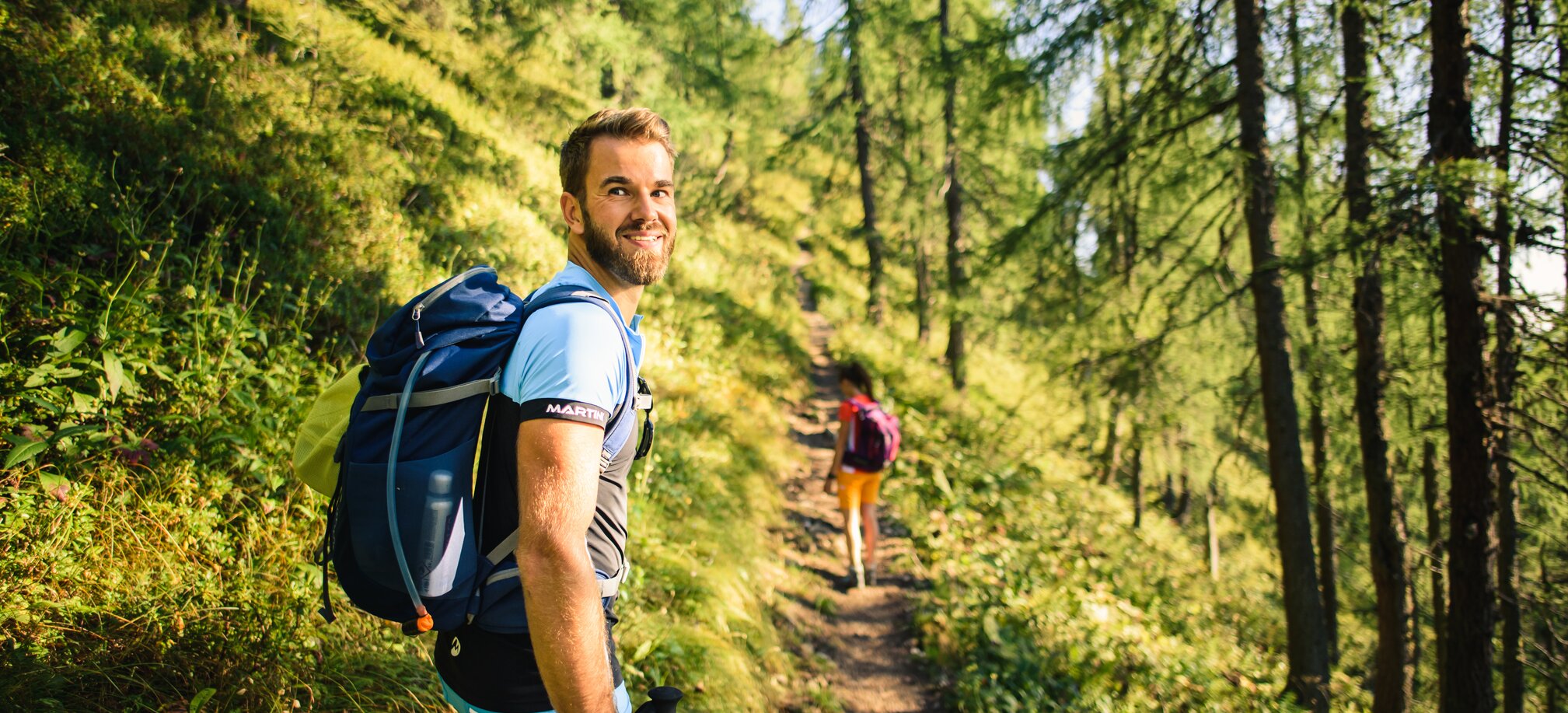 Man with backpack hikes through sunlit alpine forest trail, smiling back at camera; woman walking ahead in the background | © Salzburger Sportwelt - Christian Schartner