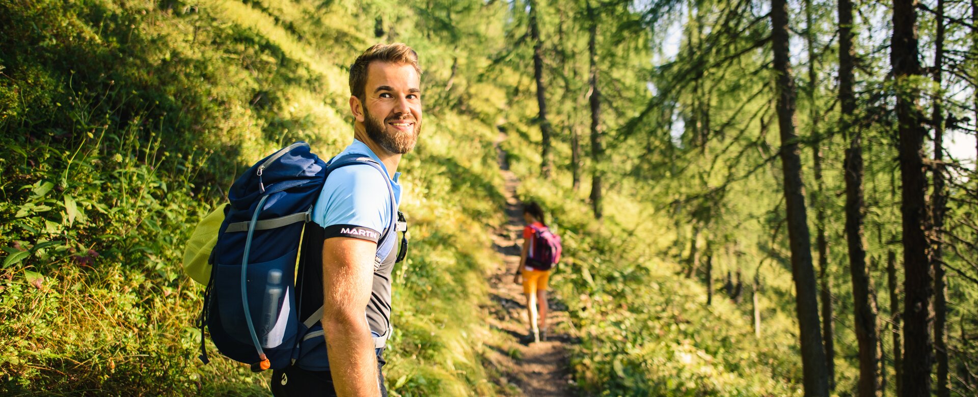 Man with backpack hikes through sunlit alpine forest trail, smiling back at camera; woman walking ahead in the background | © Salzburger Sportwelt - Christian Schartner