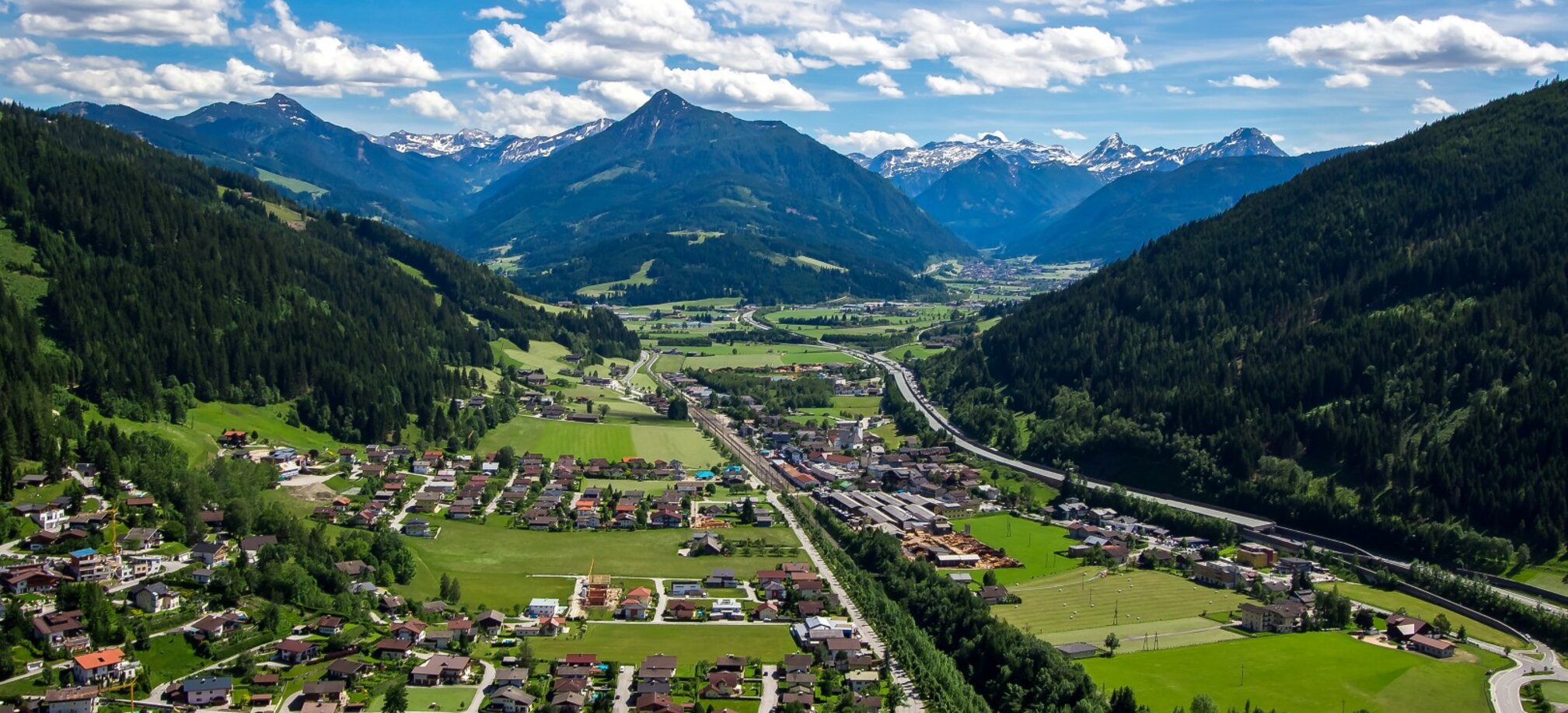 Panoramic view of Eben im Pongau with homes, fields and highway, nestled in alpine valley under a sunny summer sky