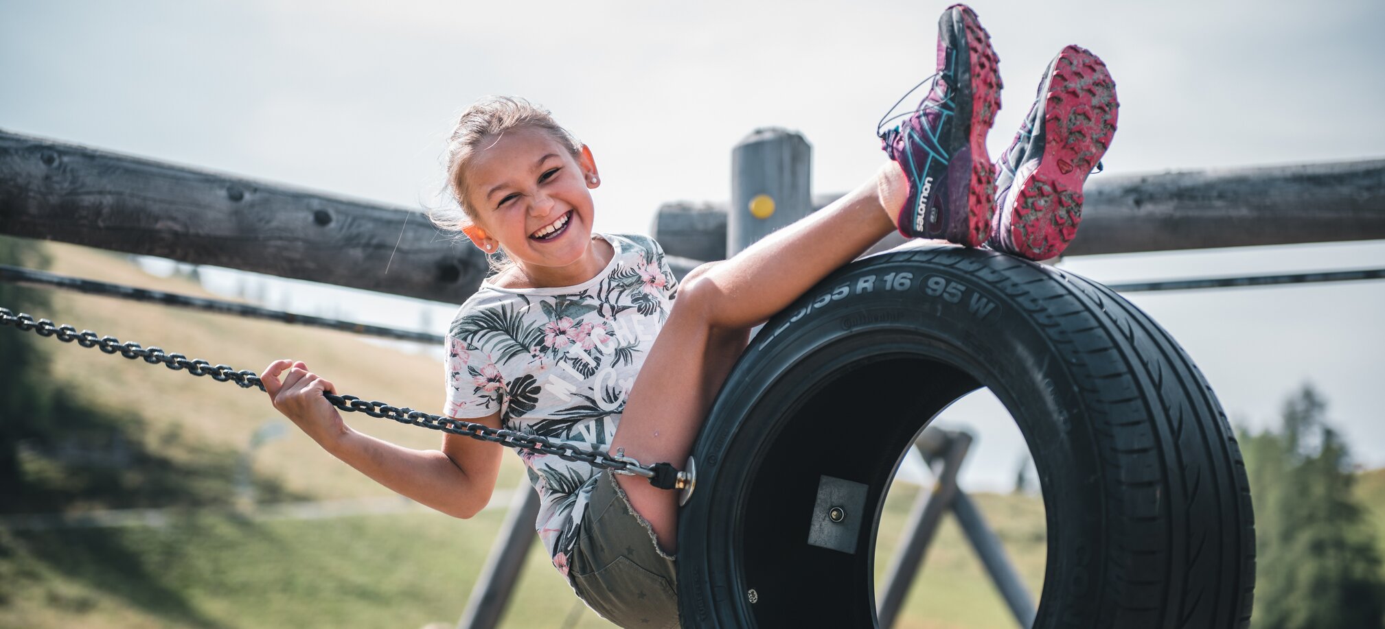 Smiling girl sits on a tire swing at the playground, holding onto chains, with grassy summer landscape in the background | © Salzburger Sportwelt