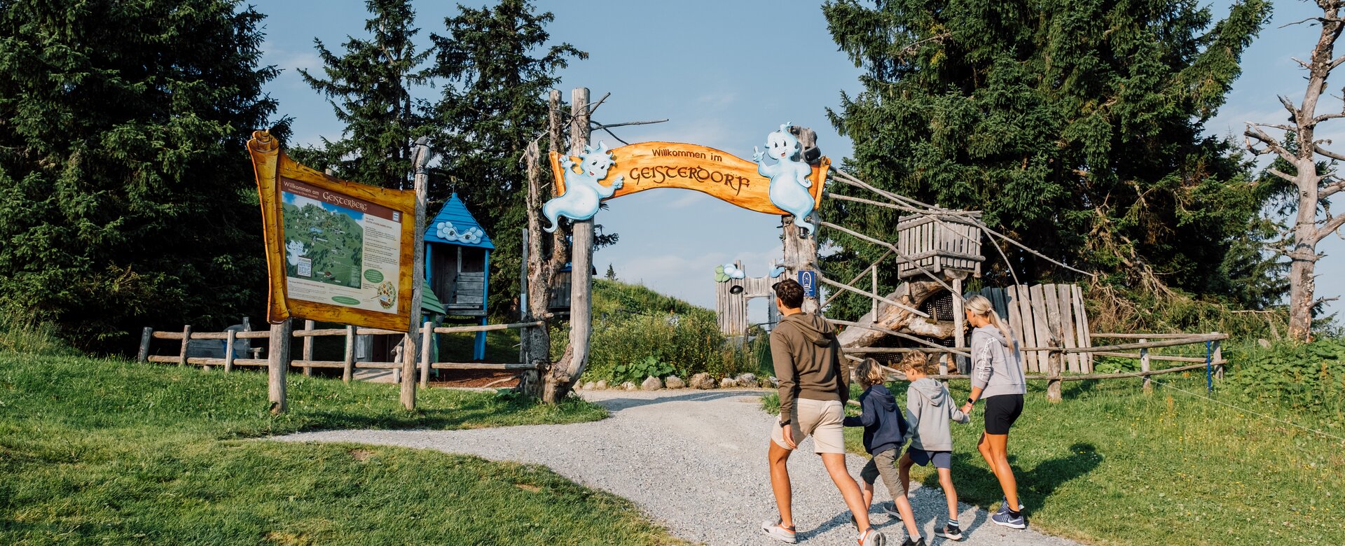 Family with two kids enters the playful wooden gateway to Geisterberg under sunshine, surrounded by trees and ghost figures