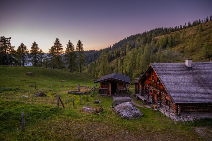 A rustic wooden hut stands on the side and is framed by an alpine meadow and trees as the sun has just set. | © Christian Schartner