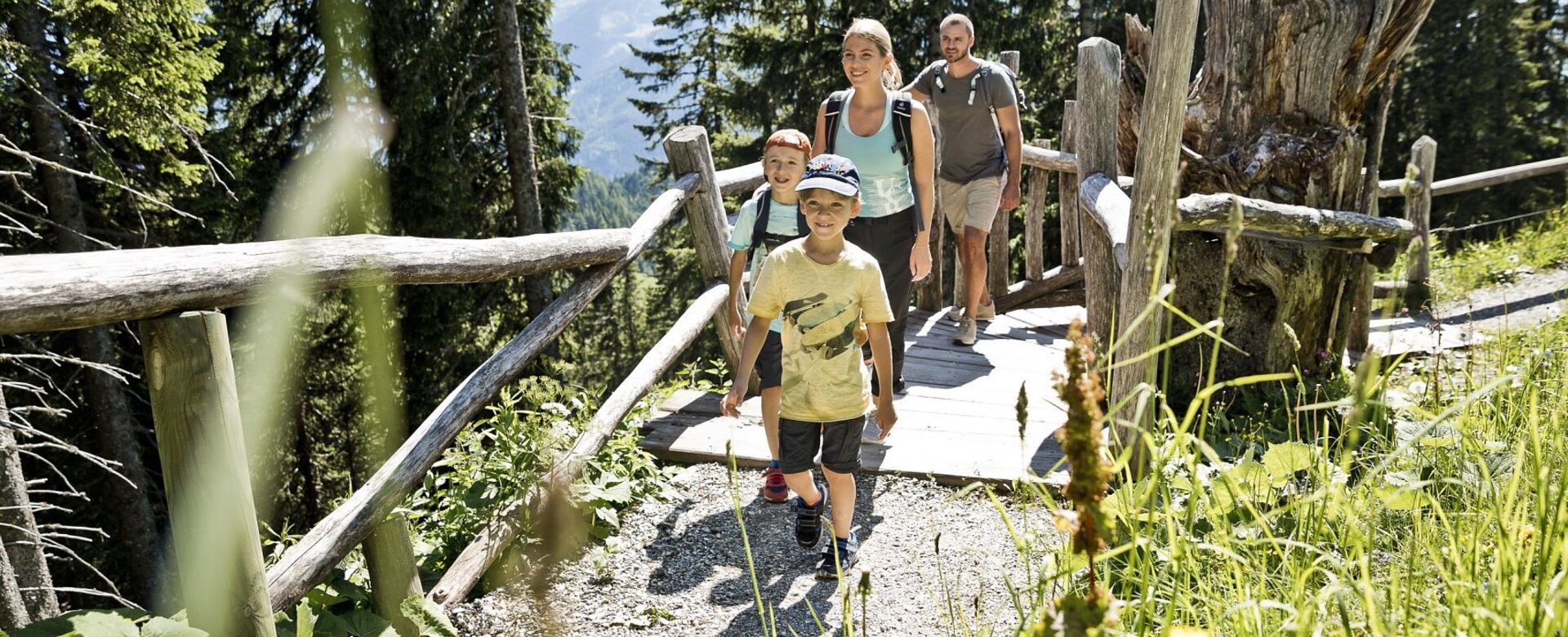 A family with two children hikes along a narrow forest trail with a wooden railing. Sunshine and lush greenery surround them. | © Salzburger Sportwelt/MirjaGeh