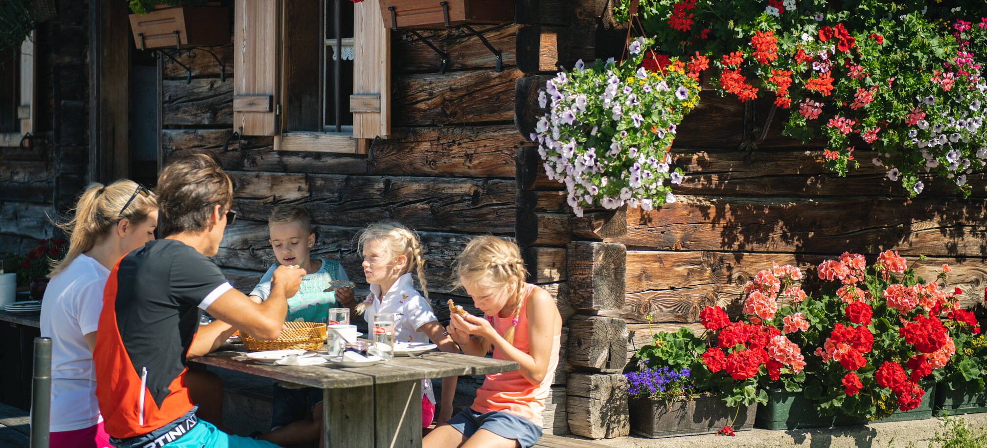 A family enjoys a snack at a wooden table in front of a rustic alpine hut decorated with colorful flowers in Filzmoos. | © Salzburger Sportwelt/Christian Schartner