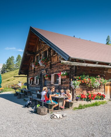 A family sits at a wooden table in front of a flower-adorned alpine hut in Filzmoos, surrounded by mountains and green meadows. | © Salzburger Sportwelt/Christian Schartner
