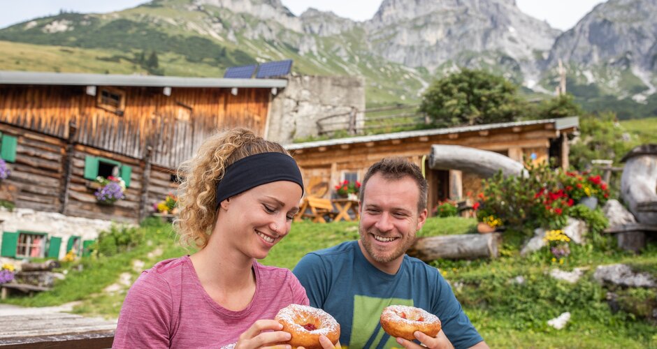 A smiling couple enjoys traditional doughnuts at a wooden table in front of an alpine hut, surrounded by colorful flowers and mountains. | © Salzburger Sportwelt - Michael Grössinger