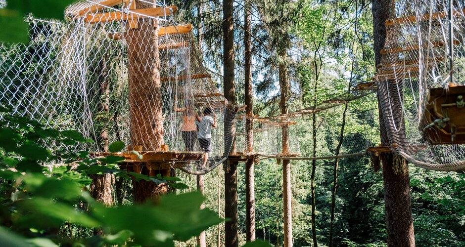Two people walk through a high ropes course with hanging bridges and platforms among tall trees. | © JOSalzburg