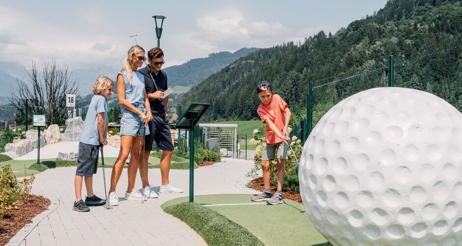 A family plays minigolf beside a large golf ball sculpture with mountains and forested slopes behind. | © JOSalzburg