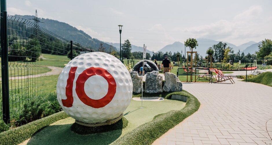 Mini golf hole with giant golf ball, flag, rocks and paths in front of meadows, mountains and blue sky | © JOSalzburg