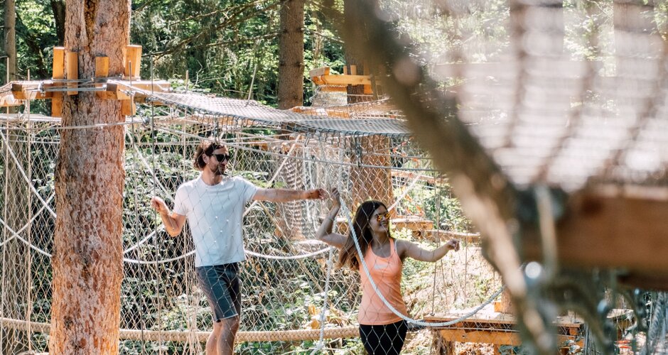 Two people balance on a rope in a high ropes course between platforms and nets in the forest. | © JOSalzburg