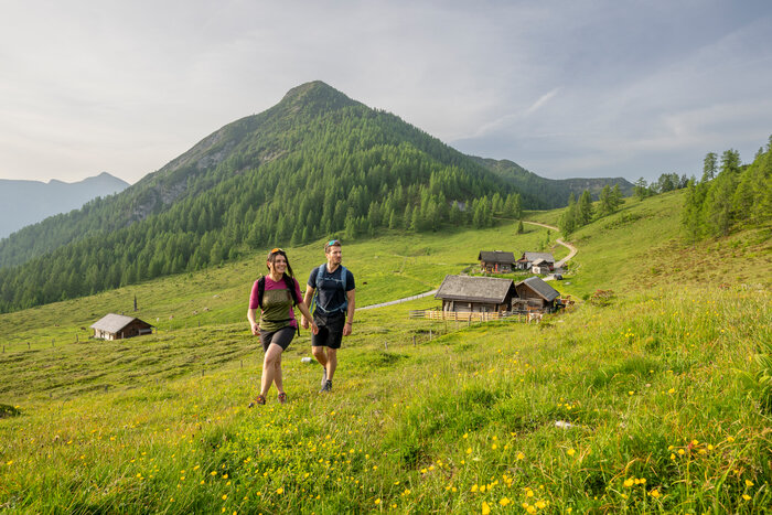 Two hikers walk across an alpine meadow near Altenmarkt, with wooden huts and a forested mountain. | © Lorenz Masser